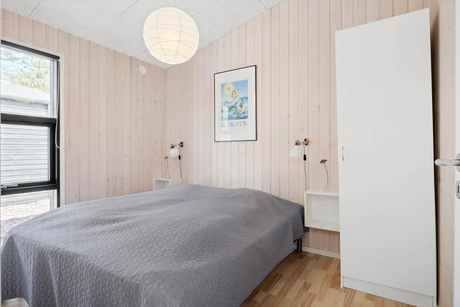 Schlafzimmer mit Bett, Wandbild und Schrank. Holzwand und Fenster sichtbar.