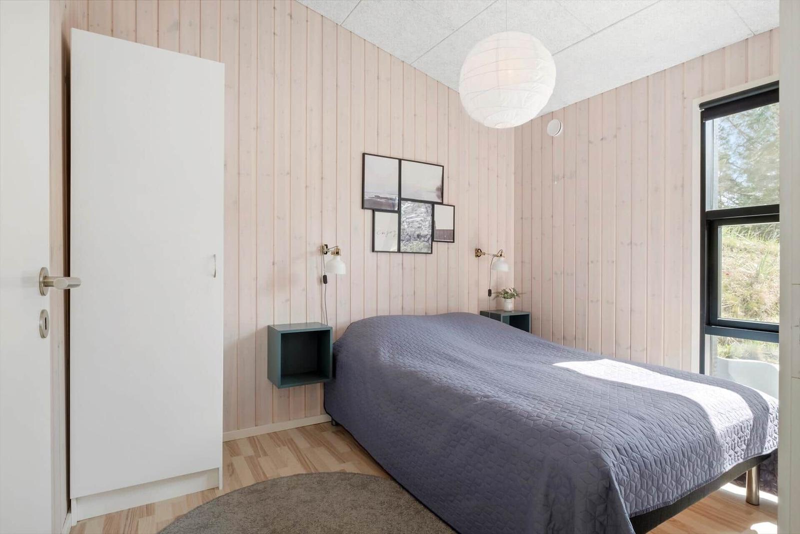 Schlafzimmer mit Bett, Wandregalen und Fenster. Holzwand und Deckenleuchte sichtbar.