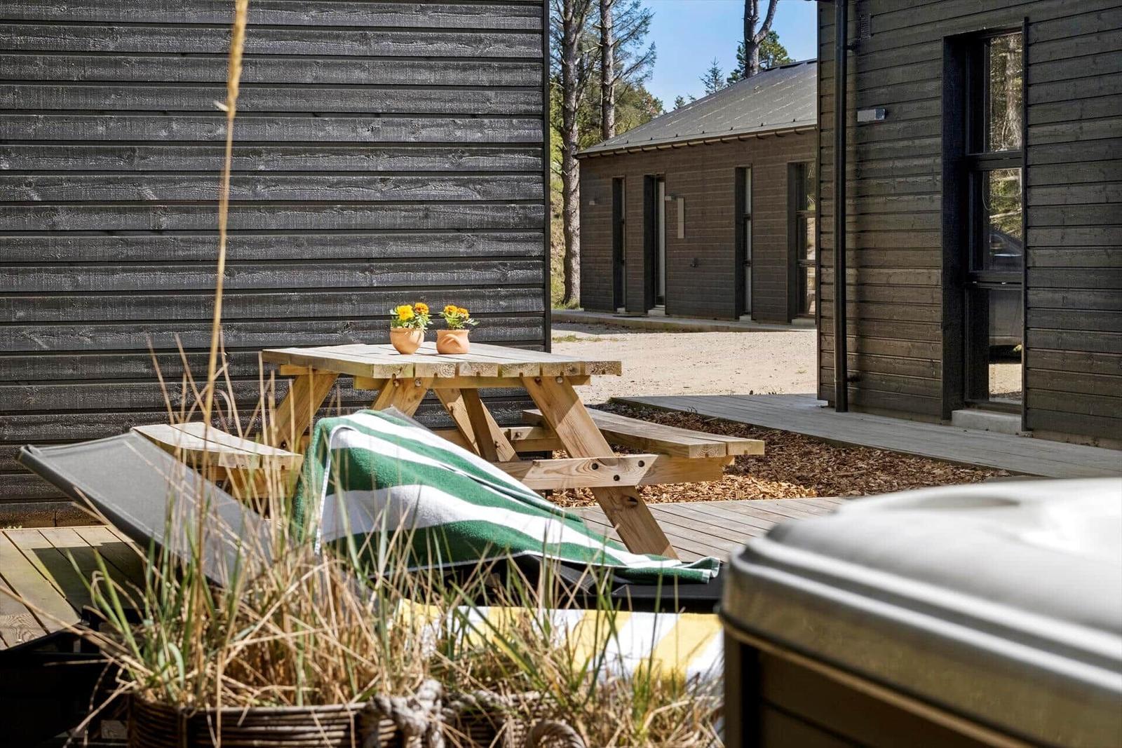 Holztisch mit Blumen, Liegestuhl und Whirlpool auf Terrasse neben Holzhütte.