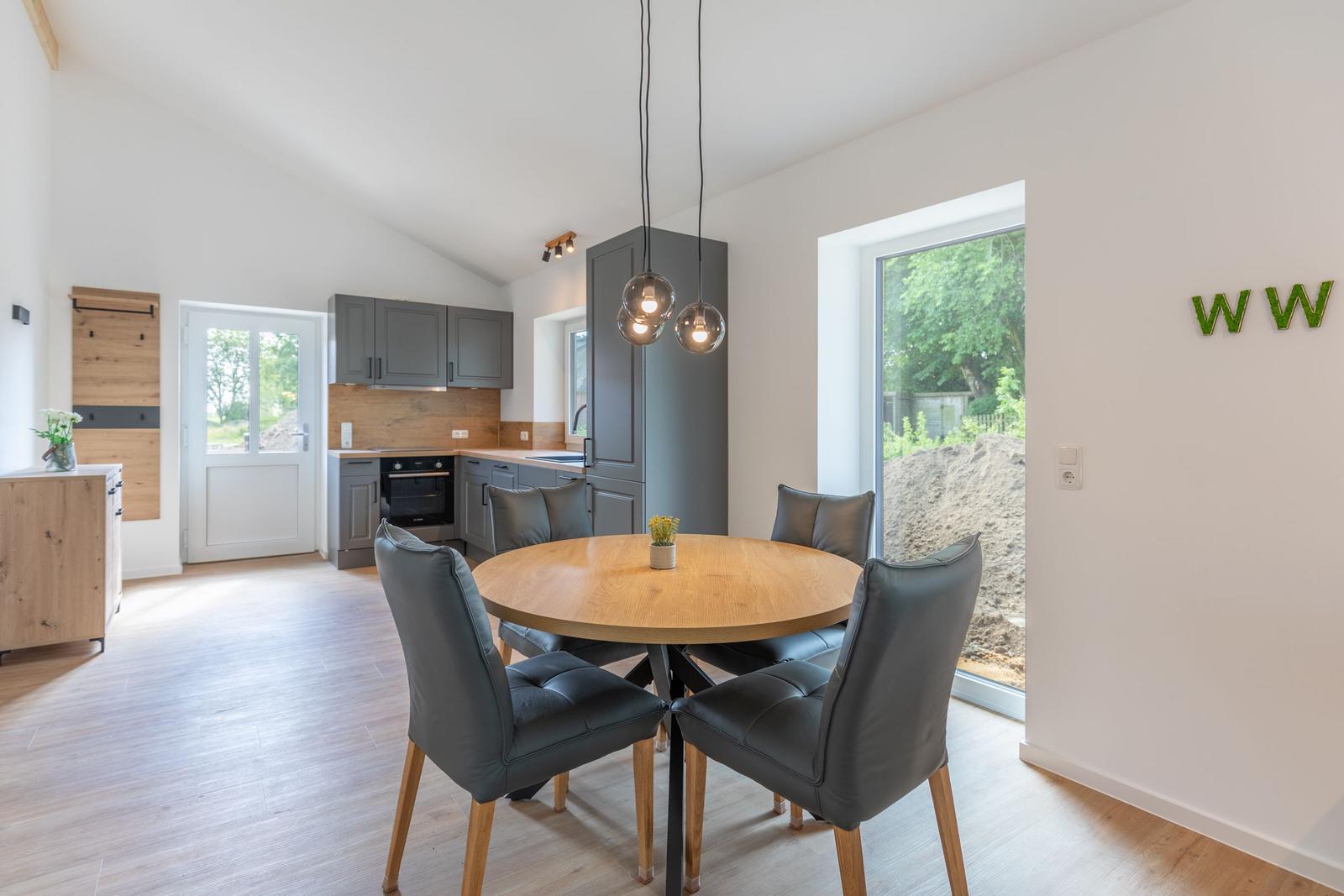 Essbereich mit runder Tisch und Stühlen. Küche mit grauen Schränken. Glastür zum Garten.