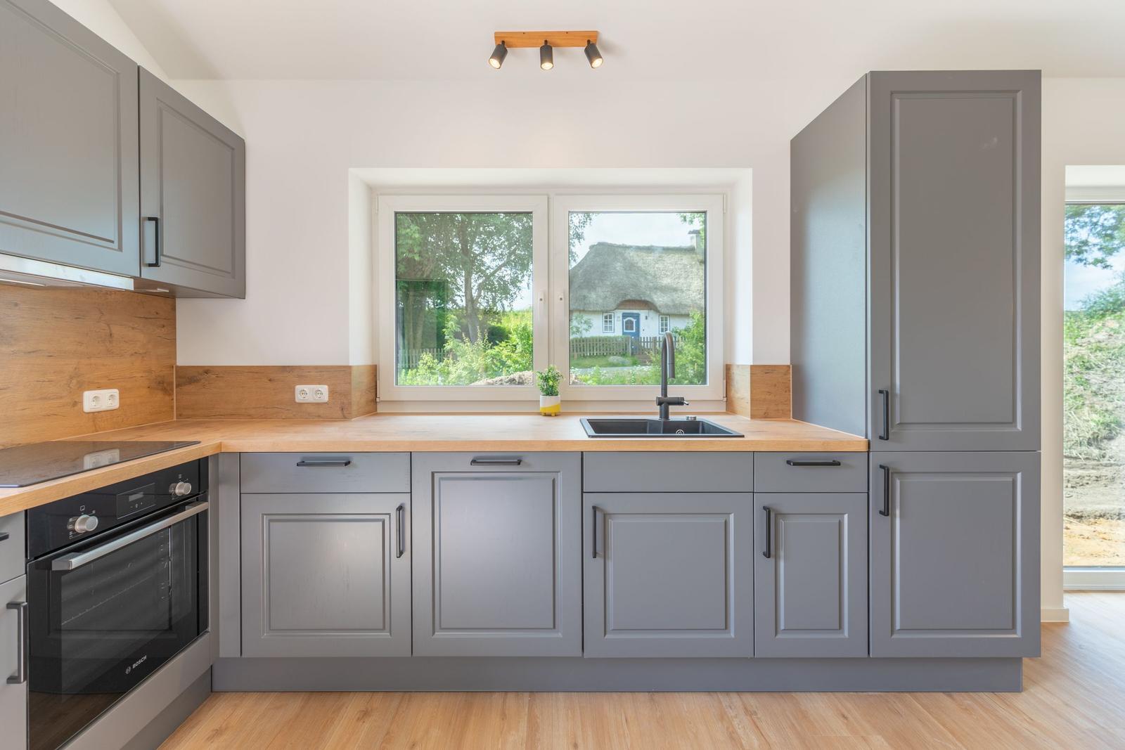 Küche mit grauen Schränken, Holz Arbeitsplatte und Fensterblick auf eine Hütte.