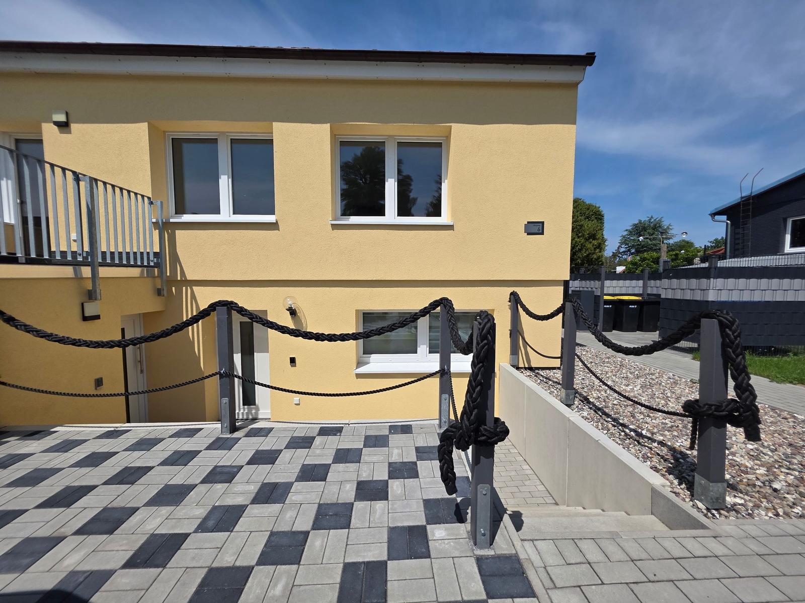 Gelbes Haus mit Balkon und schwarz-weißem Kiesweg. Vorhof mit Seilgeländer.