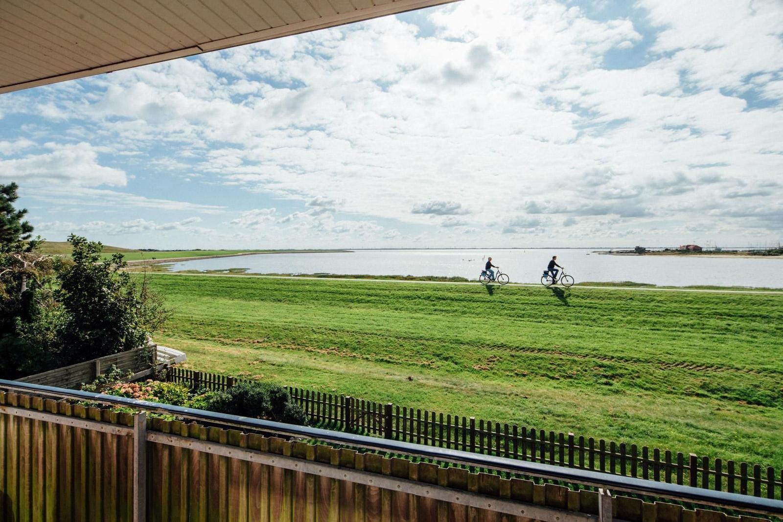 Von der Terrasse aus Blick auf grünes Feld und Wasser mit zwei Radfahrern.