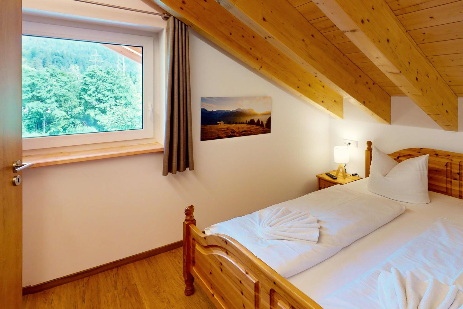 Schlafzimmer mit Holzbett, Fensterblick auf Wald und Holzdecke mit Balken.