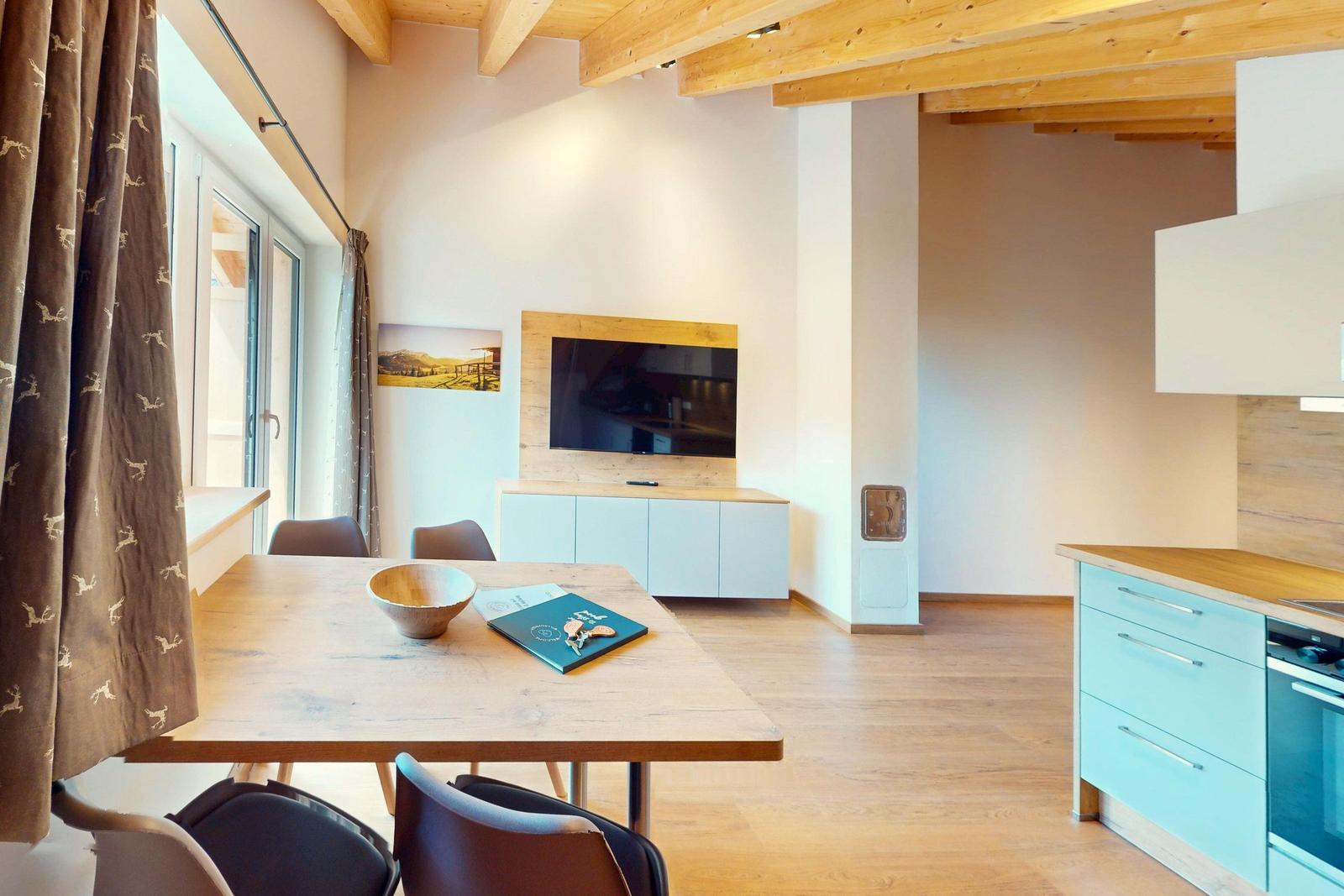 Essbereich mit Holztisch, Küche mit hellblauen Schränken und Wohnbereich mit Fernseher.
