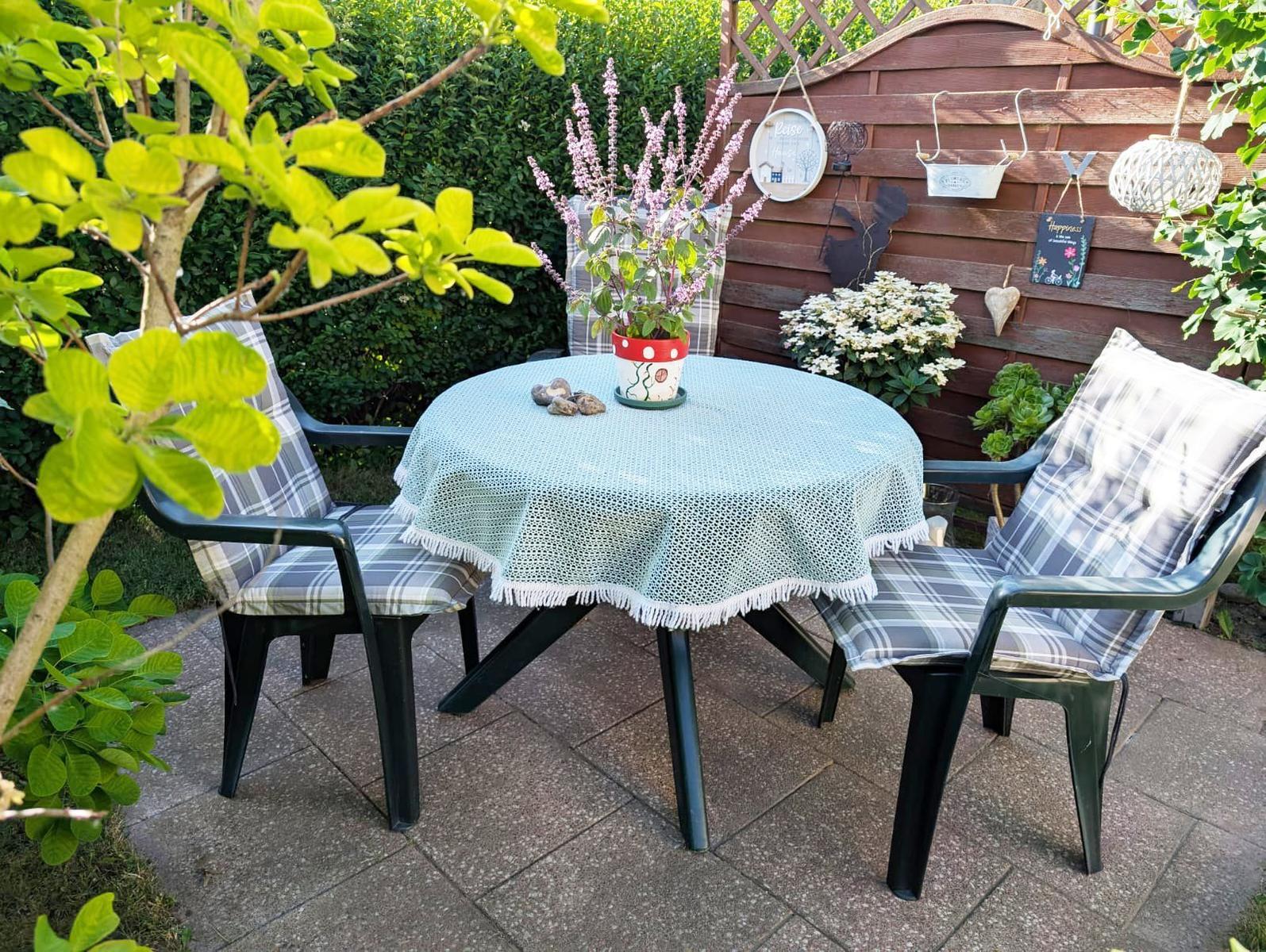 Round table with tablecloth and two chairs in the garden. Background: wooden fence and plants.