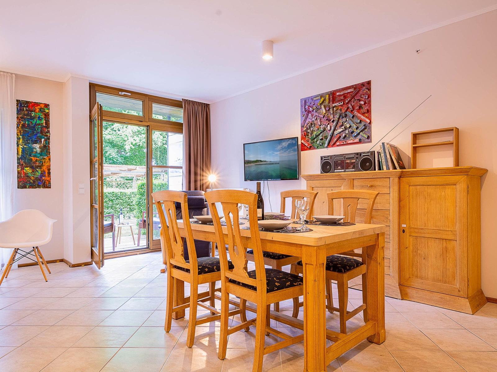 Essbereich mit Holztisch und Stühlen, Fernseher und Kabinett. Tür führt auf Balkon mit Gartenblick.