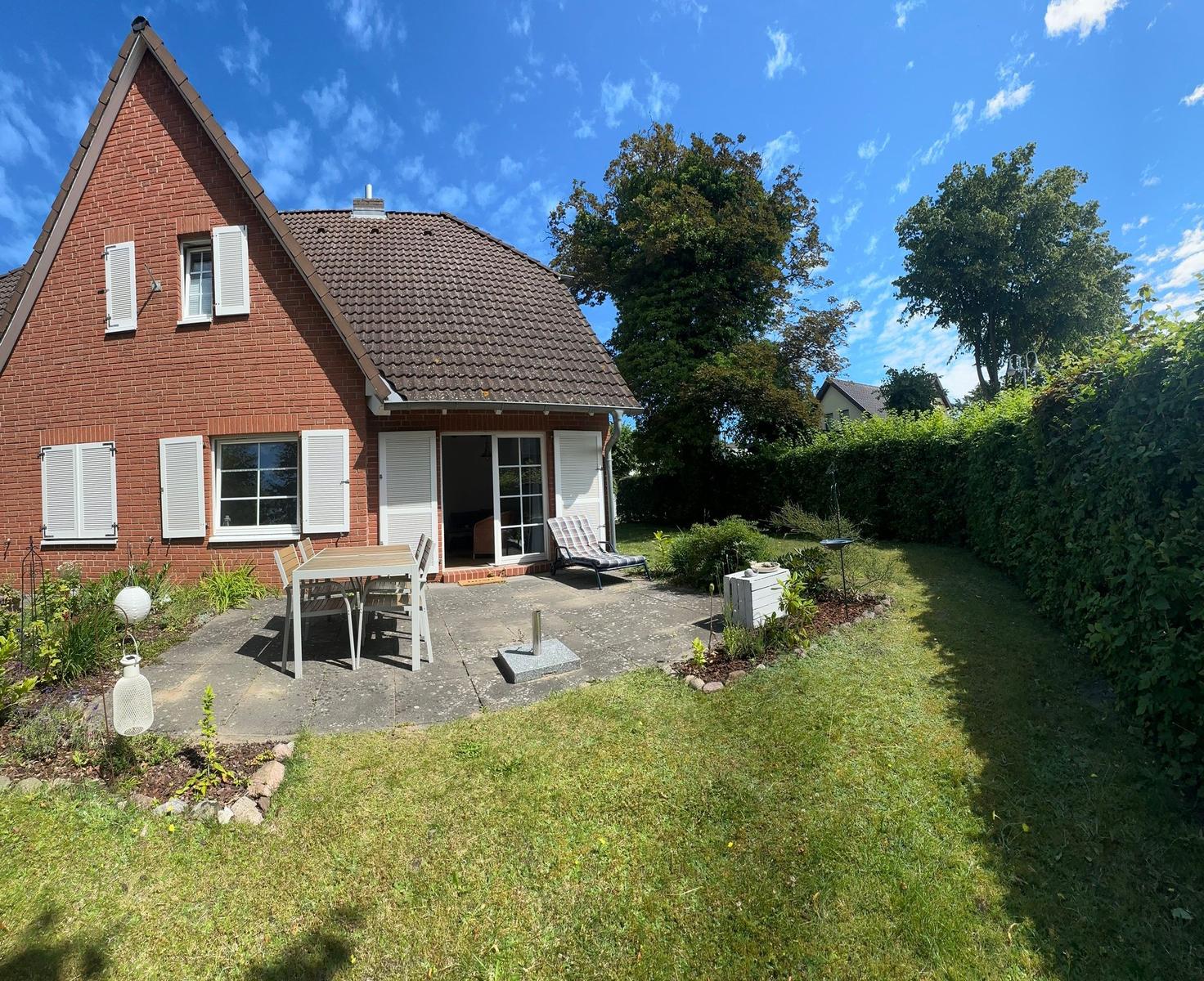 Rotes Backsteingebäude mit Terrasse, Garten und Sitzgelegenheiten unter blauem Himmel.