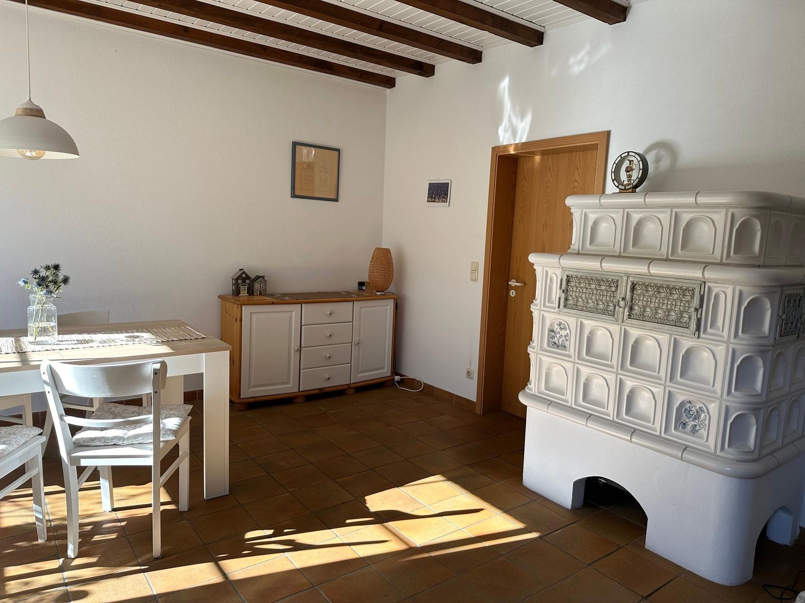 Esszimmer mit rustikalem Ofen, Holztisch und Stühlen. Deckenbalken und Fliesenboden.