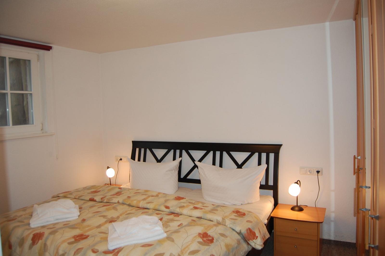 Schlafzimmer mit Bett, Nachttisch und Lampen. Fenster und Kleidung sind sichtbar.