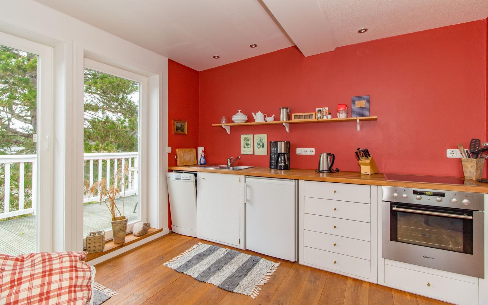 Küche mit roten Wänden, weißen Schränken und Holzoberflächen. Blick auf Balkon mit Blick auf Bäume.