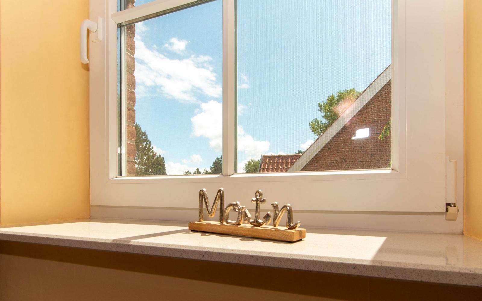 Fenster mit Blick auf Dach und Himmel, dekoratives Objekt mit Schriftzug auf Fensterbank.