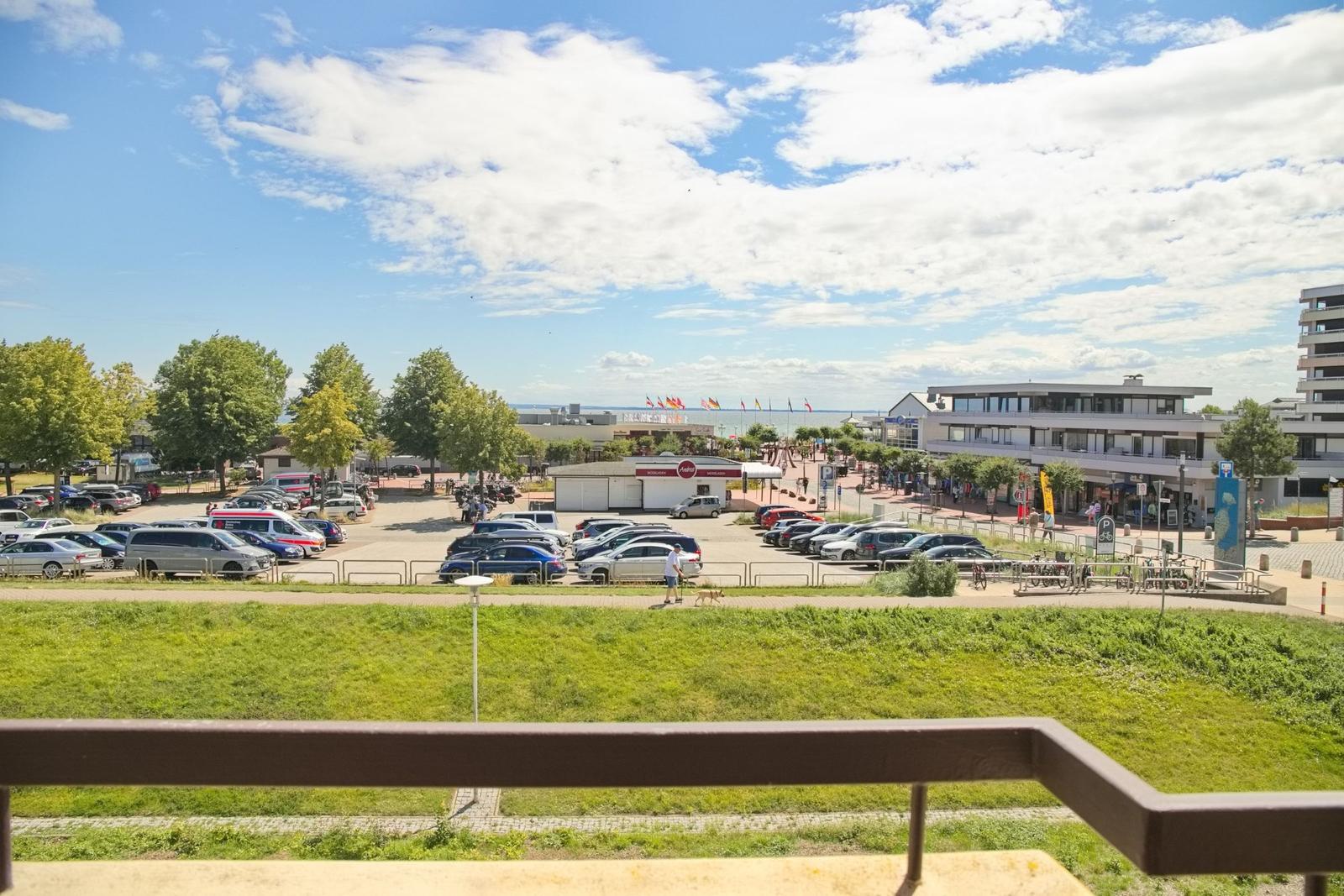 Blick auf Parkplatz, Grünfläche und Strandpromenade mit Gebäuden und Fahnen.