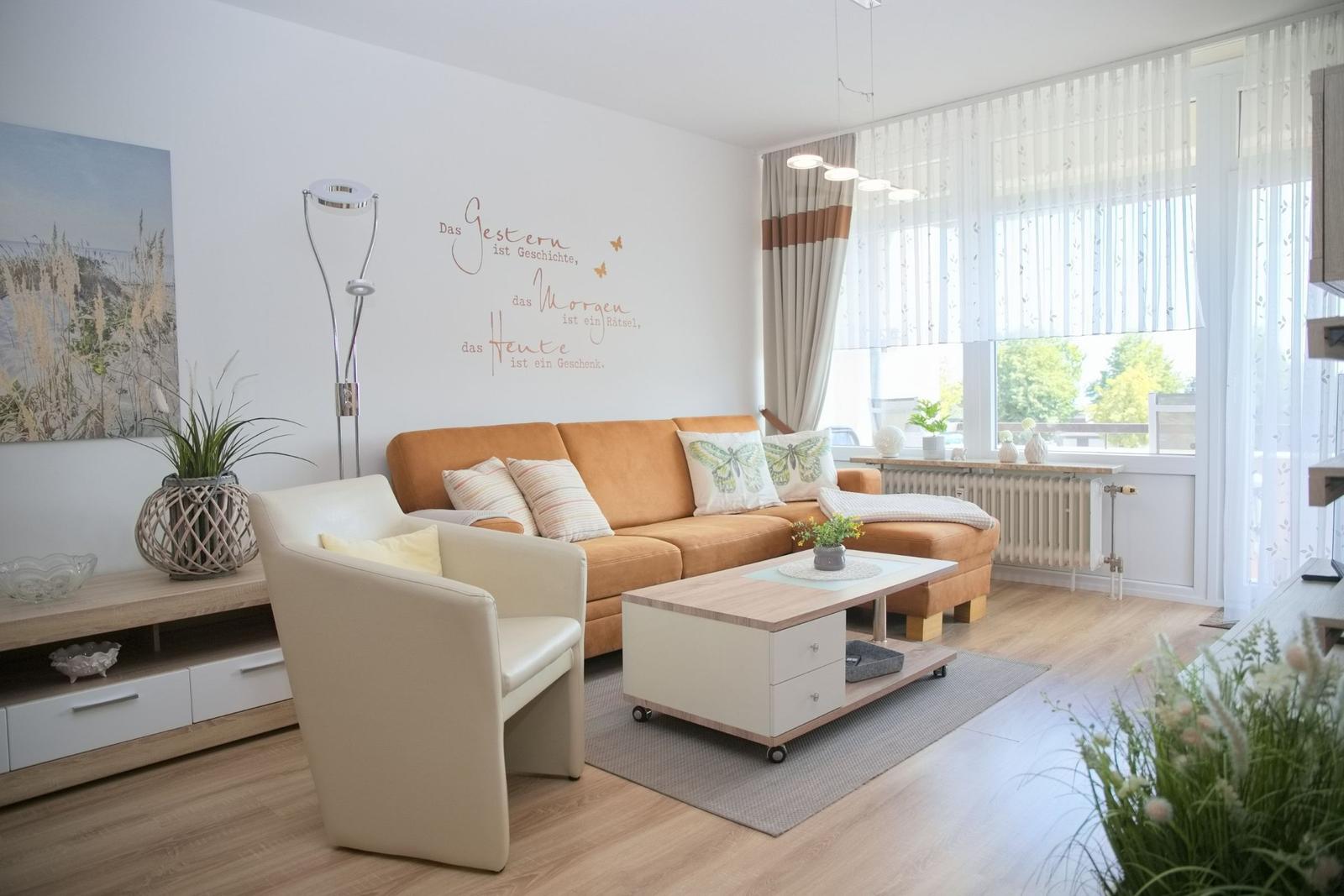 Wohnzimmer mit orange Sofa, weißen Sessel und Holzboden. Große Fenster mit Blick nach draußen.