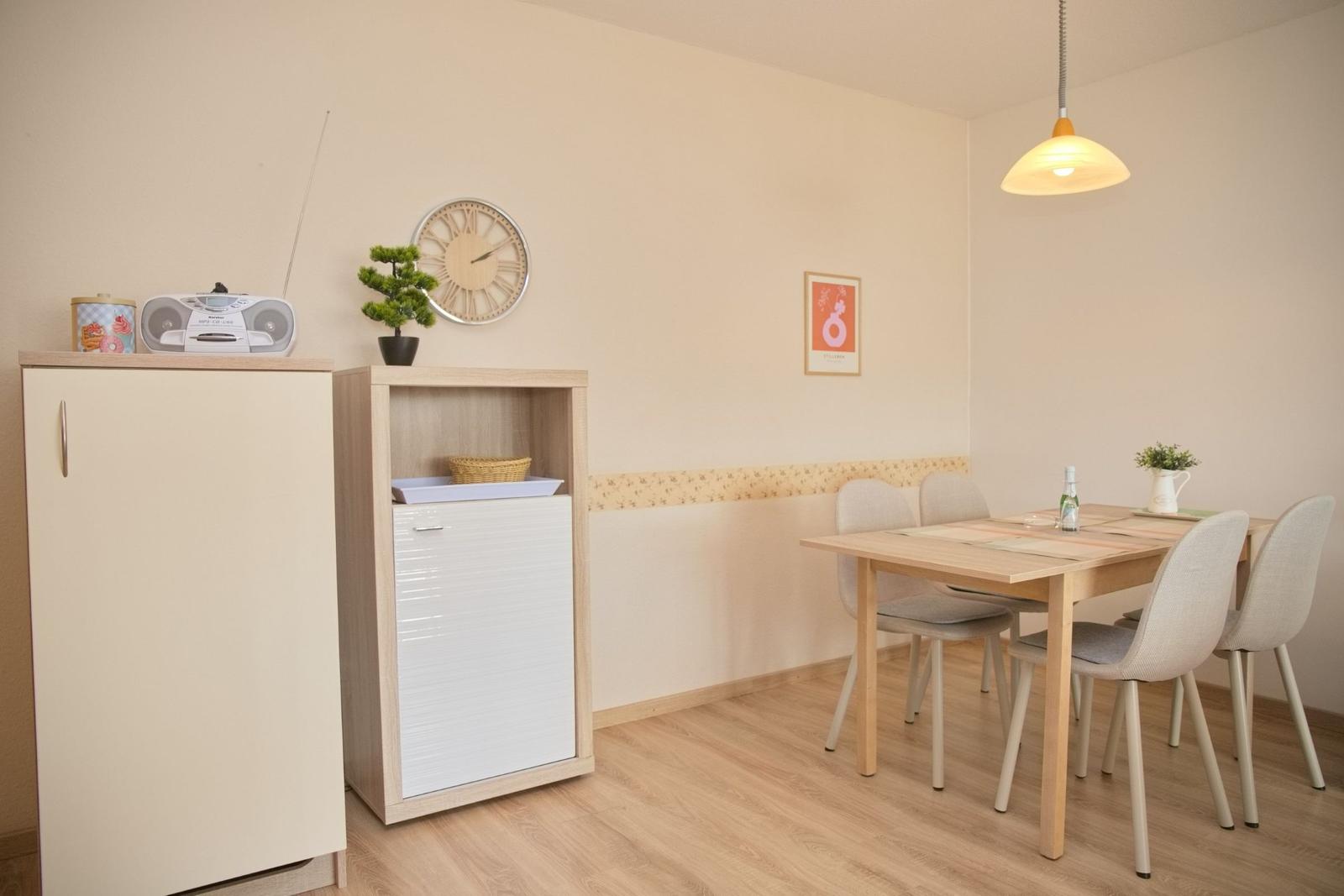 Essbereich mit Tisch, vier Stühlen und Küchenmöbeln. Wanduhr und Pendelleuchte sind sichtbar.