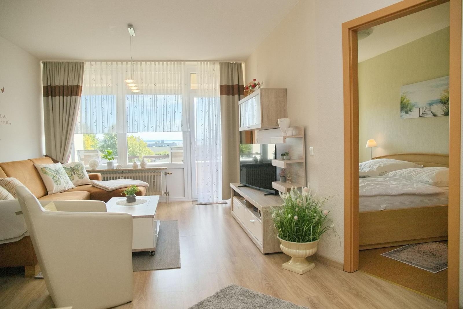 Wohnzimmer mit Sofa, Couchtisch und Fernseher. Blick auf Balkon. Schlafzimmer mit Bett sichtbar.