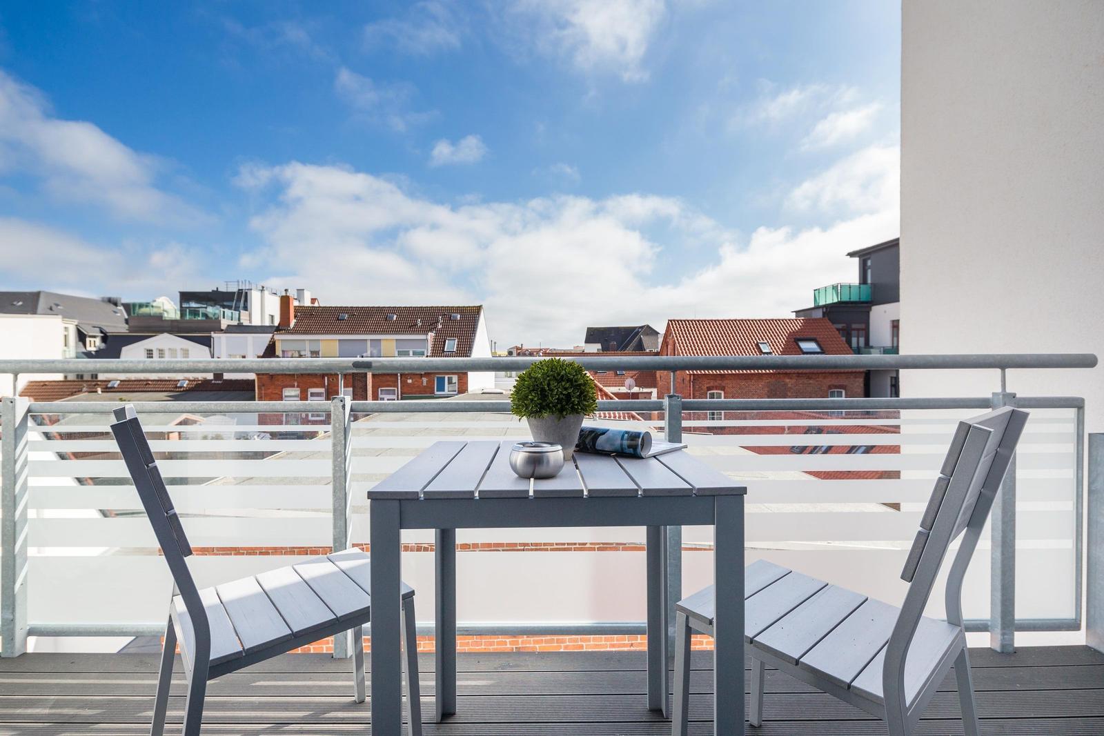 Terrasse mit Tisch und zwei Stühlen. Blick auf Dächer und Himmel.