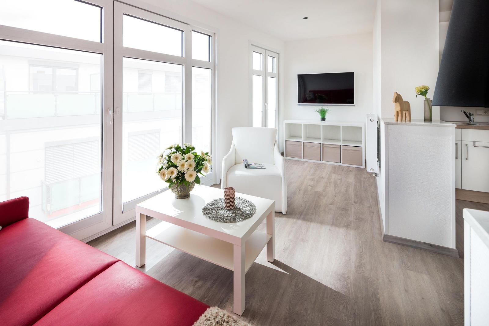 Wohnzimmer mit rotem Sofa, weißem Sessel und TV. Große Fenster und Holzboden.