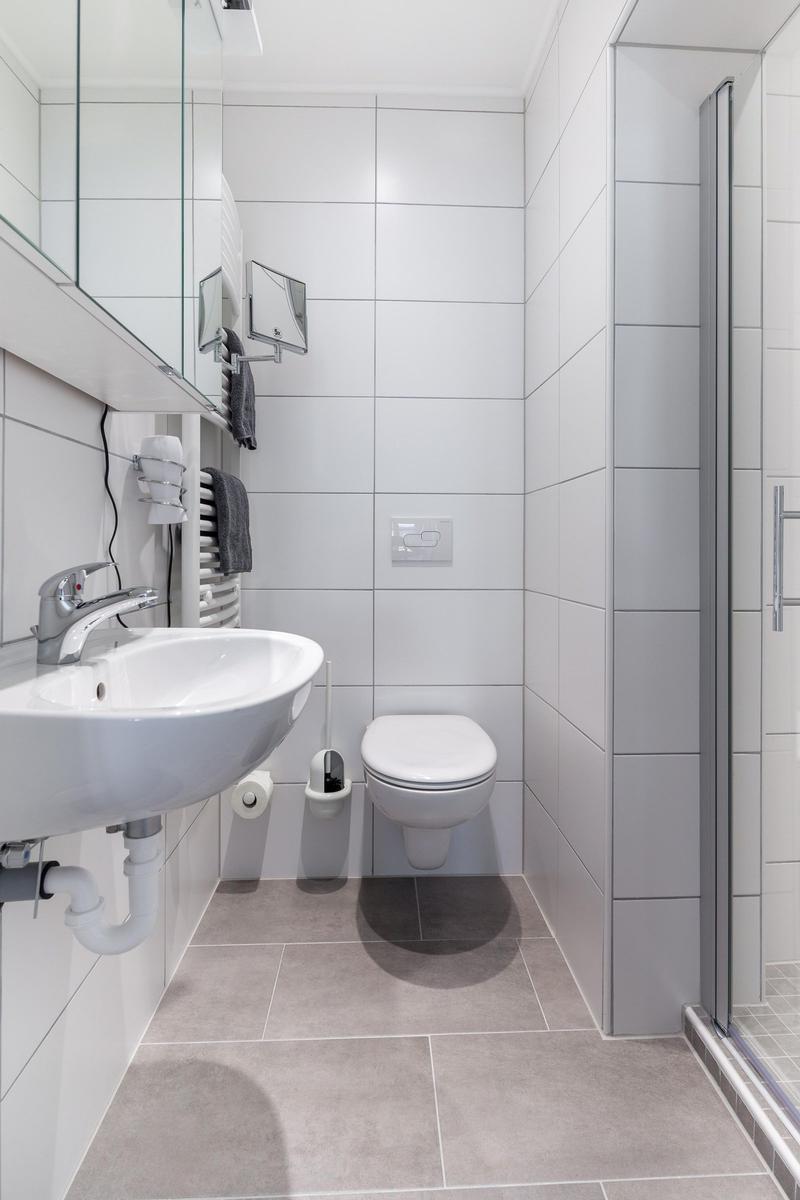 Badezimmer mit Waschbecken, Toilette und Dusche. Weiße Fliesen und graue Bodenfliesen.