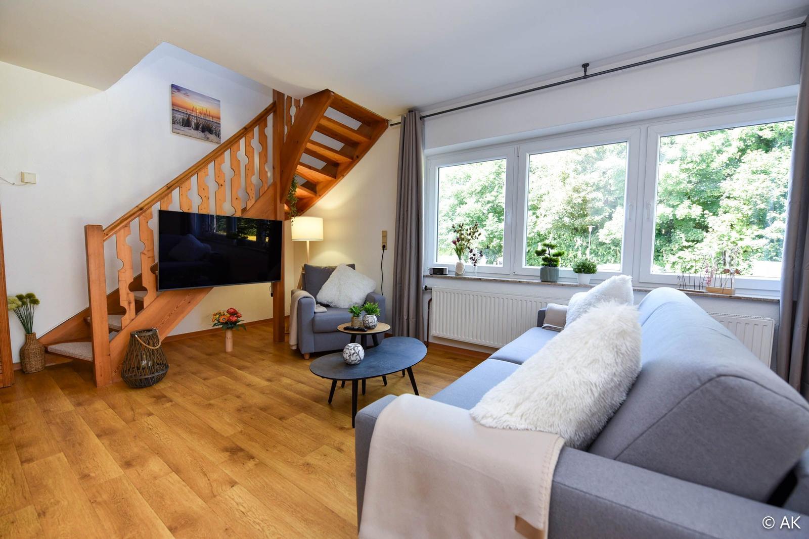 Wohnzimmer mit Sofa, Sessel und TV unter Holztreppe. Fenster mit Blick auf Bäume.