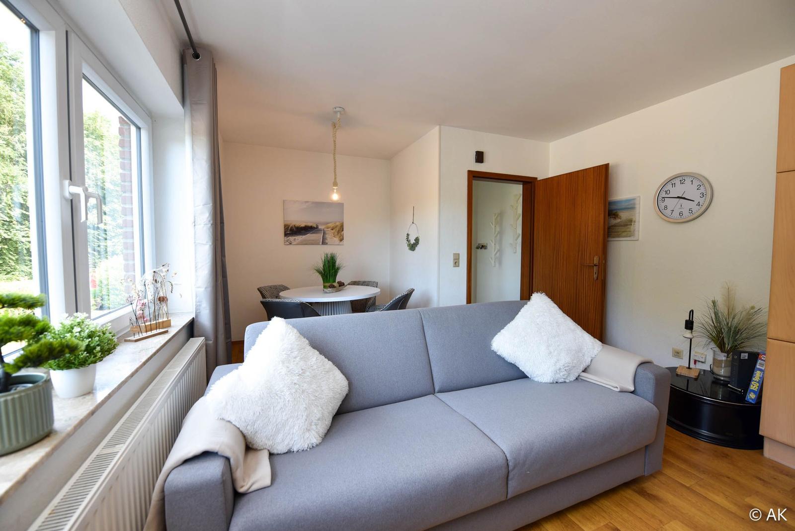 Ein Wohnzimmer mit grauem Sofa, weißen Kissen und Holzboden. An der Wand hängt eine Uhr.