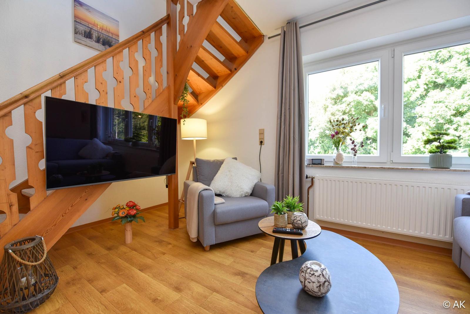 Wohnzimmer mit Holztreppe, TV und Fenster mit Blick auf Bäume.