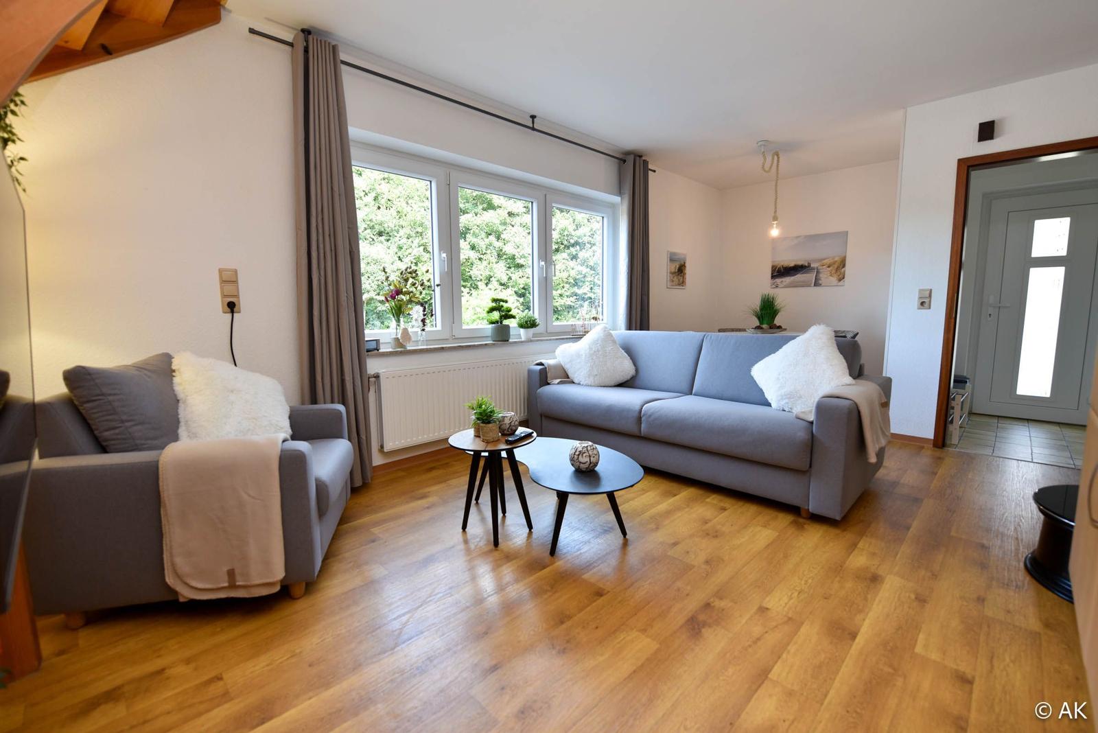 Wohnzimmer mit Sofa, Sessel und Holzboden. Fenster mit Blick auf Grün.