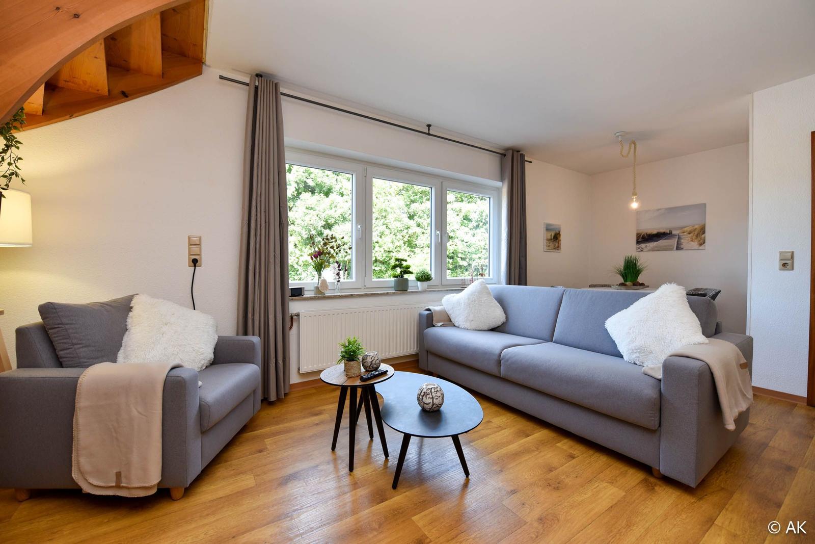 Wohnzimmer mit Sofa, Sessel und Holzboden. Fenster mit Blick auf Grün.