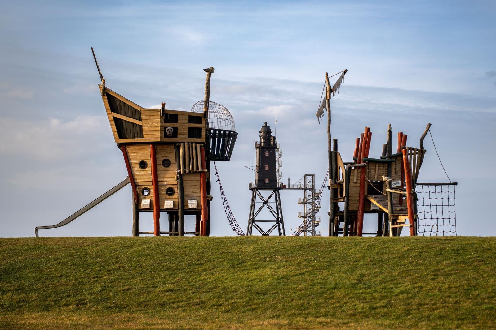 Pirate ship playground with slide and ladder on green slope. Background: black lighthouse.