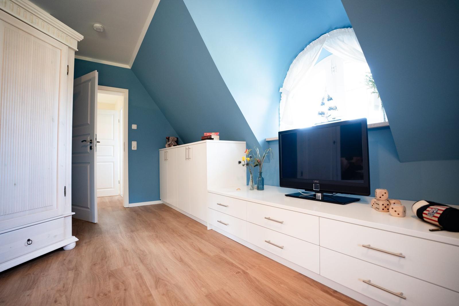 Ein Schlafzimmer mit blauen Wänden, weißem Schrank und Holzboden. TV und Fenster sind sichtbar.