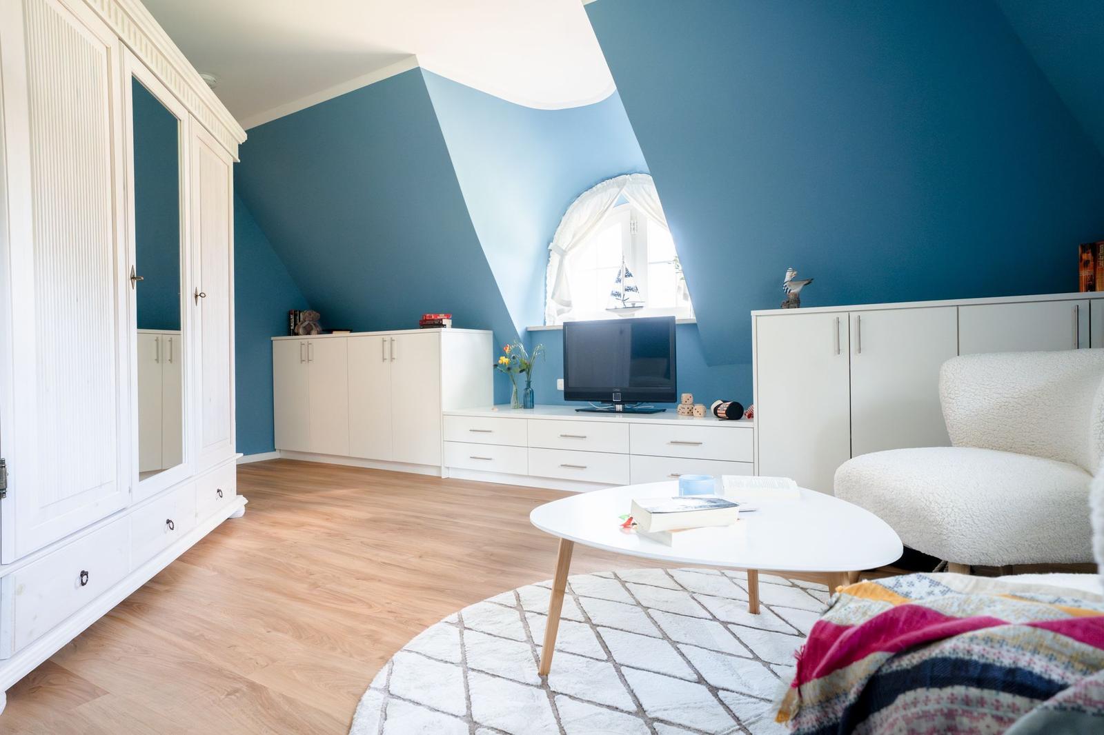 Zimmer mit blauen Wänden, weißer Einrichtung und Holzboden. Fenster und TV sichtbar.