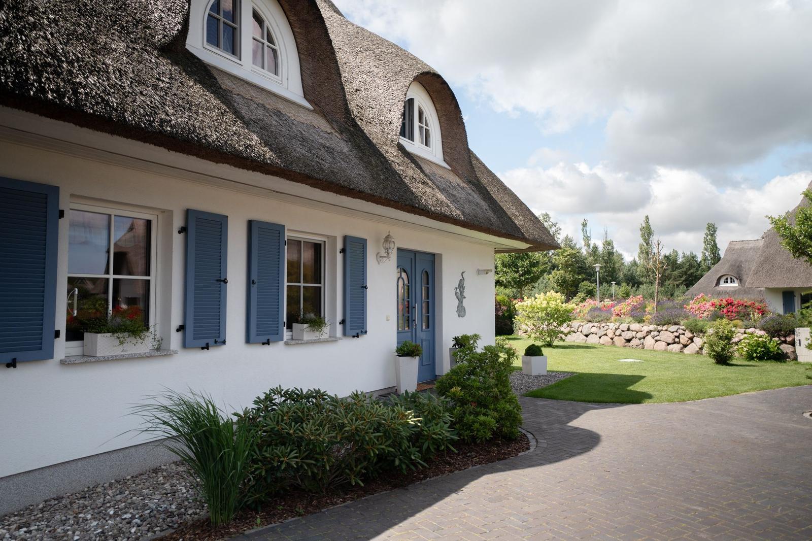 Haus mit Strohdach, blauen Fensterläden und blauer Tür. Garten mit Blumen und Rasen.