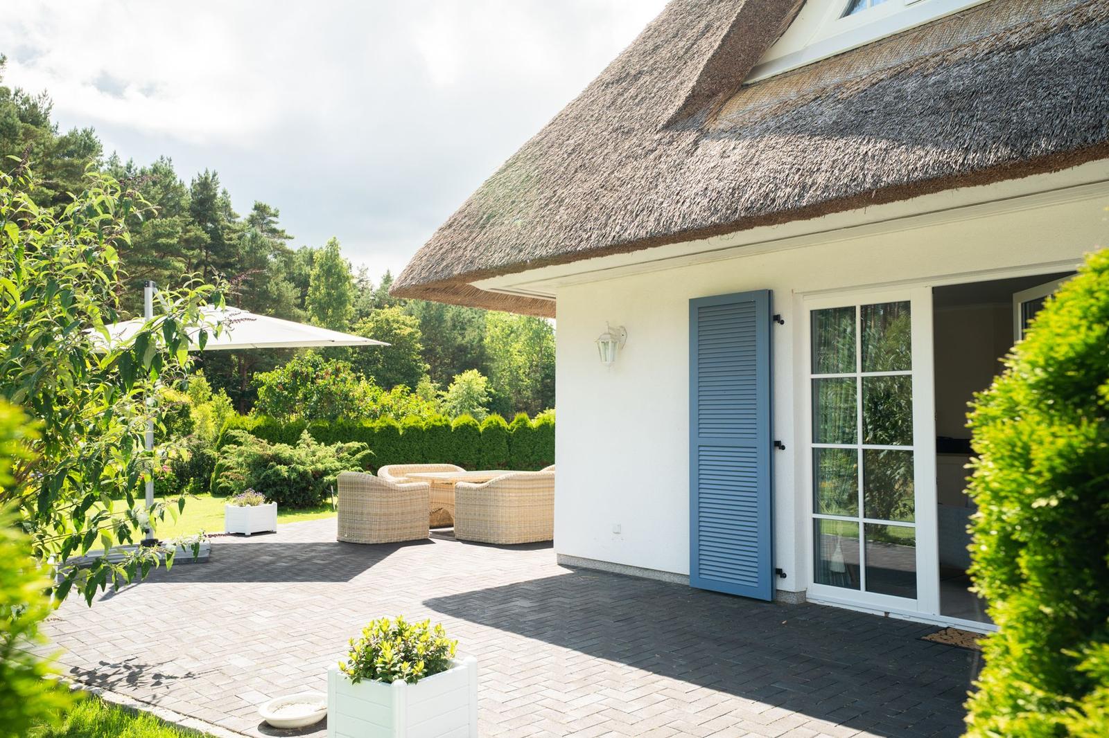 Haus mit Strohdach, Terrasse, Gartenmöbel und grüner Umgebung.