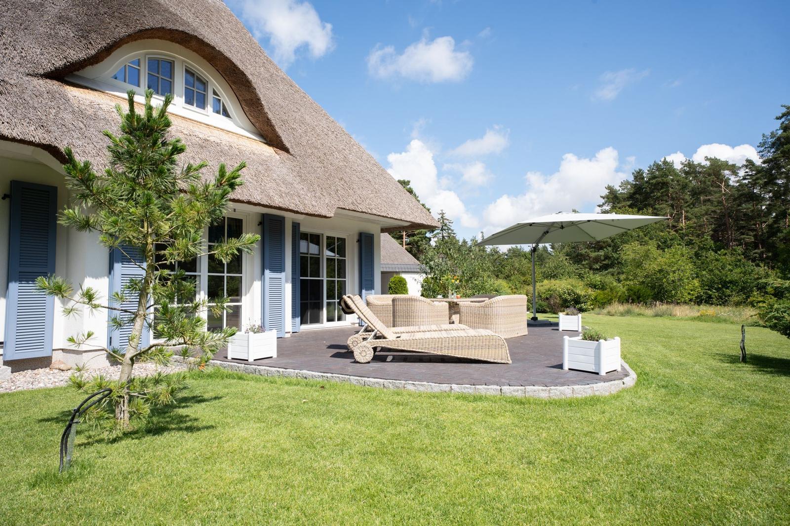 Haus mit Strohdach, Terrasse mit Liege und Sonnenschirm, umgeben von Gras und Bäumen.