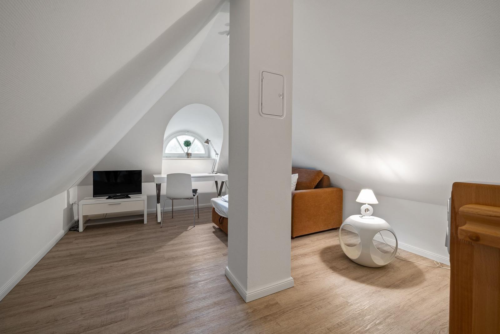 Zimmer mit Schrägdach, Sofa, TV, Schreibtisch und runden Tisch.