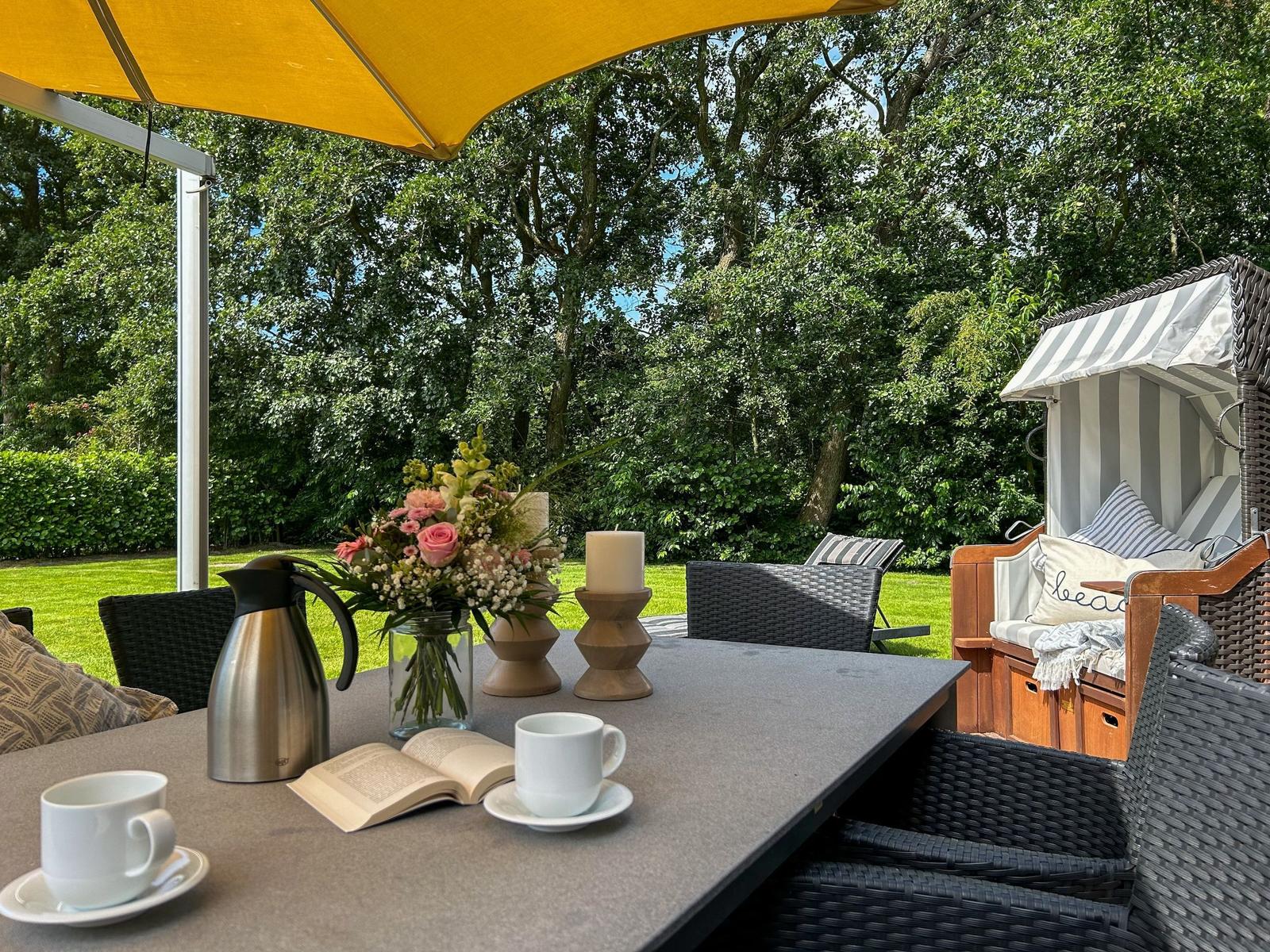 Tisch mit Kaffeetassen, Blumen und Buch unter gelbem Sonnenschirm. Strandkorb und Garten im Hintergrund.