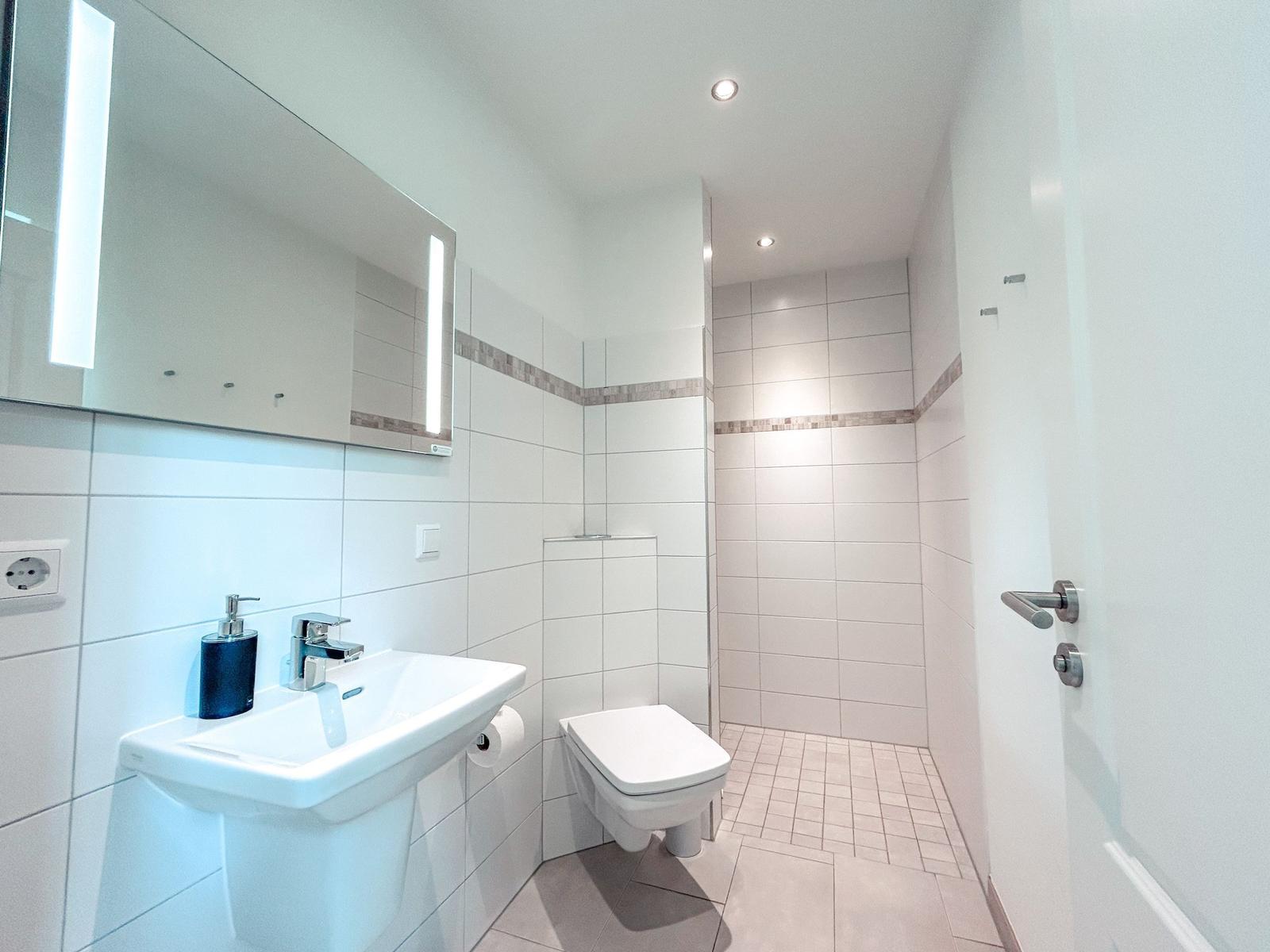 Badezimmer mit Dusche, Waschbecken und Toilette. Fliesen und Spiegel mit Beleuchtung.