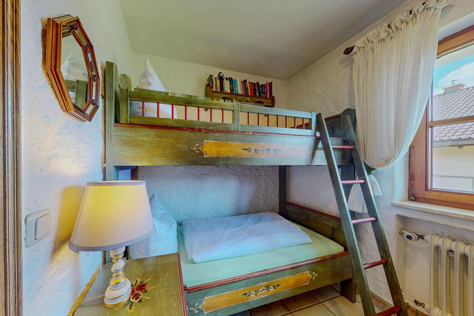 Zimmer mit Doppelstockbett, Nachttisch und Lampe. Fenster mit Vorhängen.