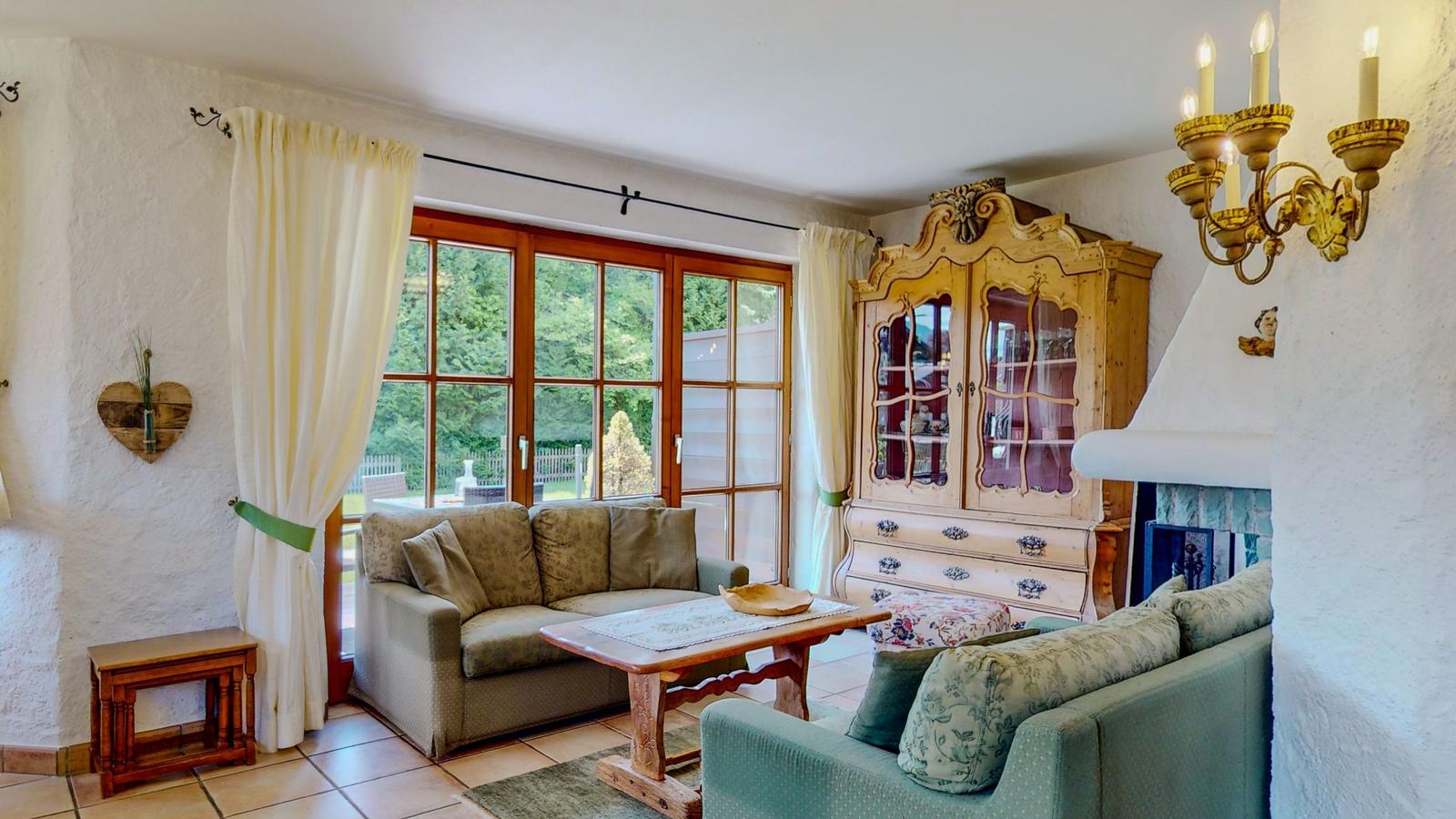 Wohnzimmer mit Sofa, Couchtisch und antikem Schrank. Große Fenster mit Blick auf Garten.