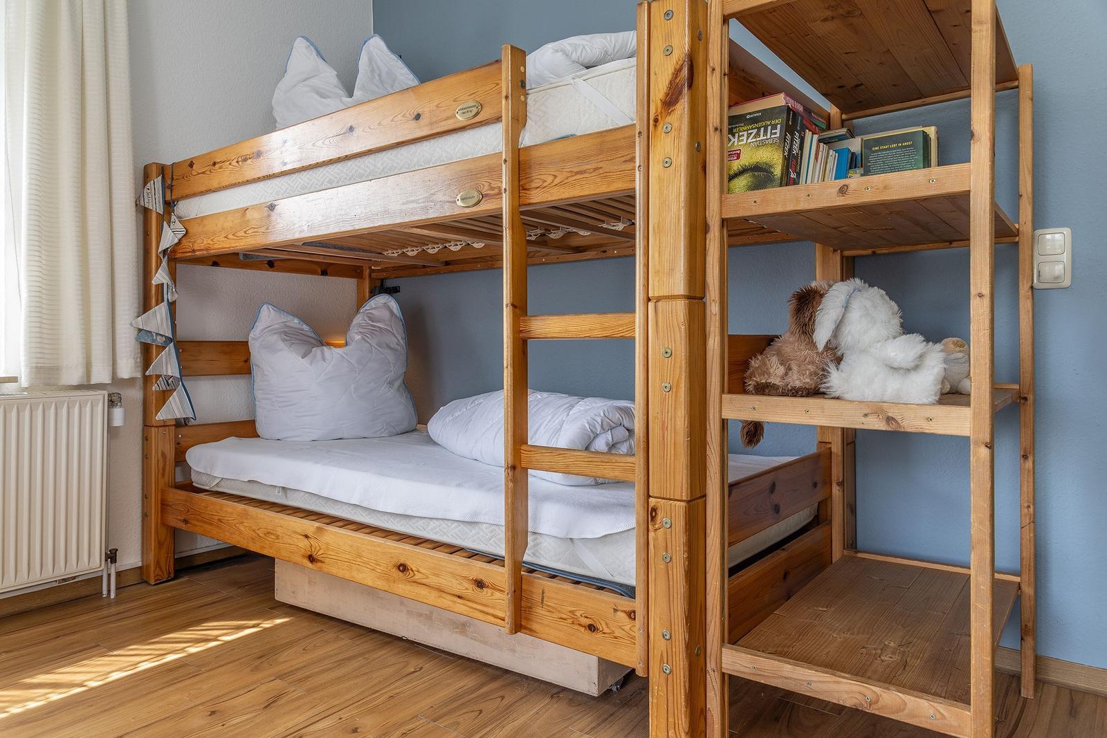 Holz-Etagenbett mit Matratzen, Kissen und Stofftieren. Bücherregal und Heizung sind vorhanden.