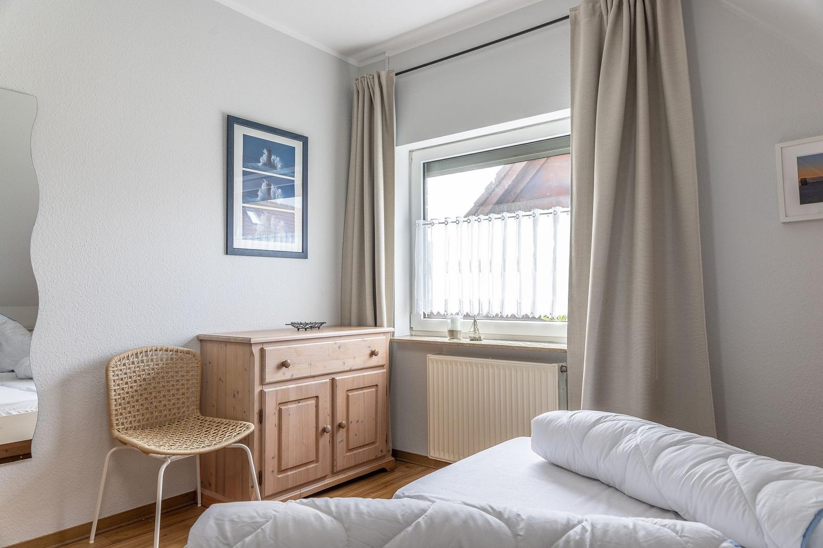 Schlafzimmer mit Bett, Holzschrank und Stuhl. Fenster mit Vorhängen und Bildern an der Wand.