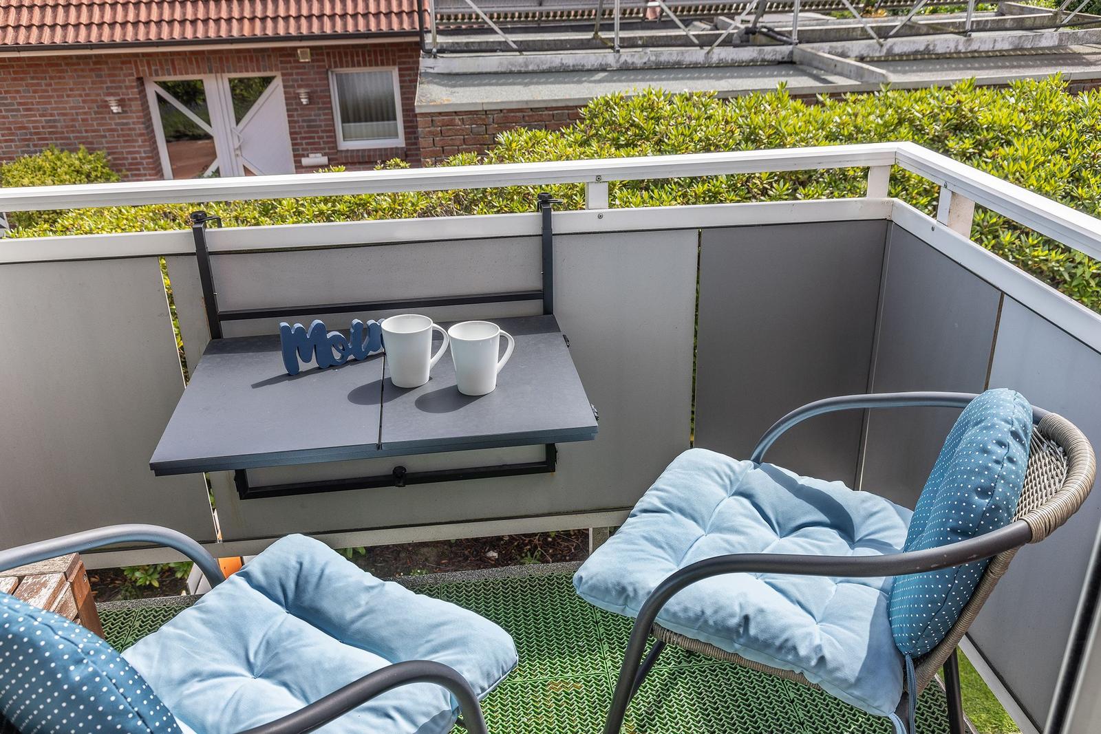 Terrasse mit Tisch, zwei Stühlen und Kissen. Hintergrund: grüne Hecke und Haus.