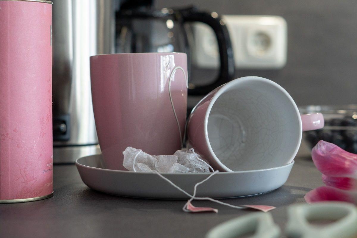 Auf der Arbeitsfläche stehen zwei Tassen, eine davon umgekippt, mit Teebeuteln und einem rosa Behälter.