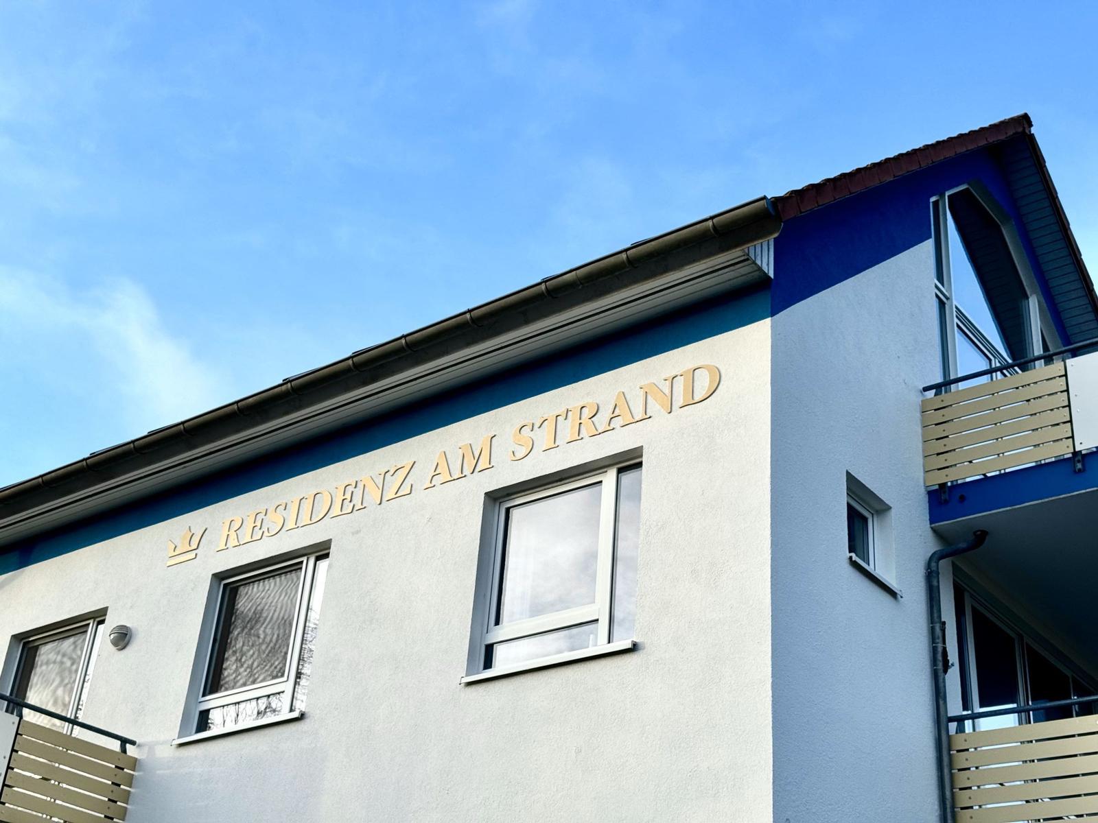 Ferienhaus mit Schild RESIDENZ AM STRAND und Balkonen unter blauem Himmel.