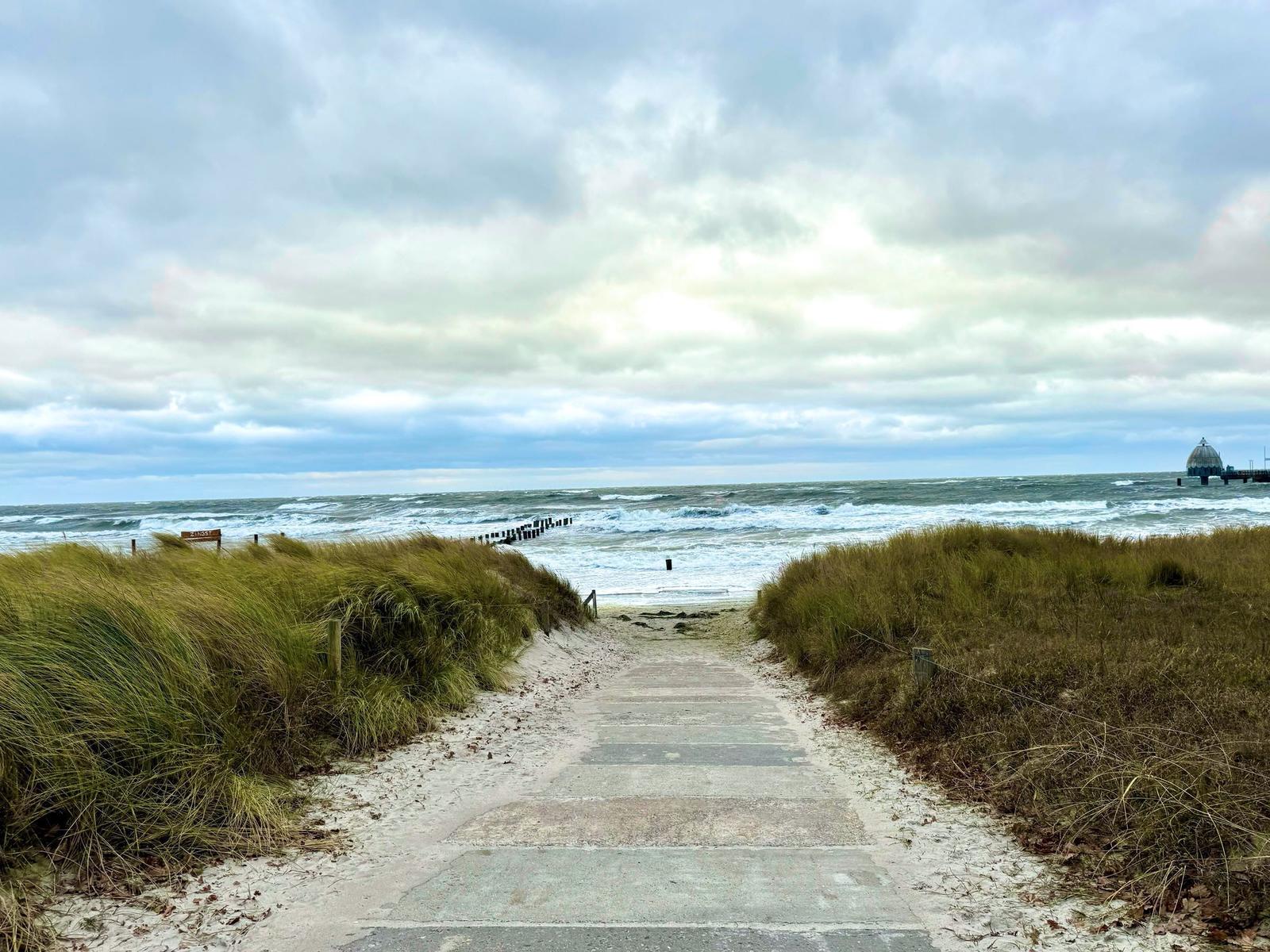 Pfad durch Dünen zur Strandpromenade mit Leuchtturm im Hintergrund.
