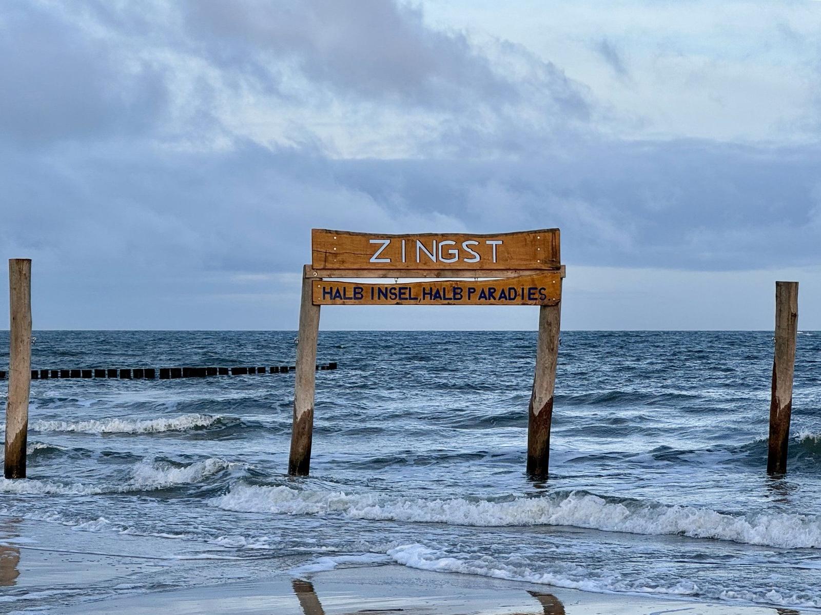 Holzschild Zingst am Strand mit Wellen im Hintergrund.