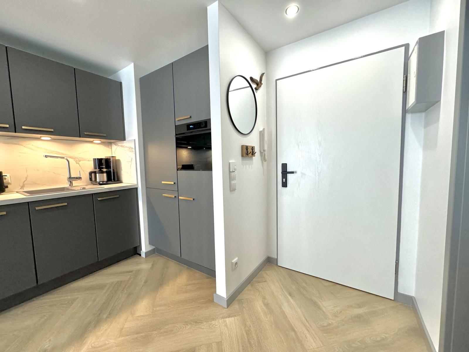 Küche mit grauen Schränken und Holzboden. Hinter dem Eingang steht ein runder Spiegel.