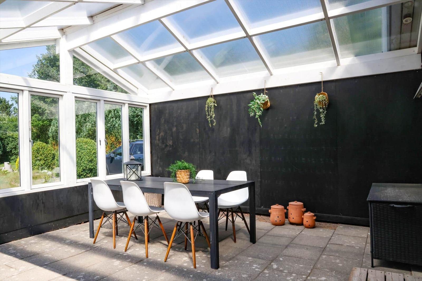 Terrasse mit Tisch, Stühlen und Pflanzen unter Glasdach.