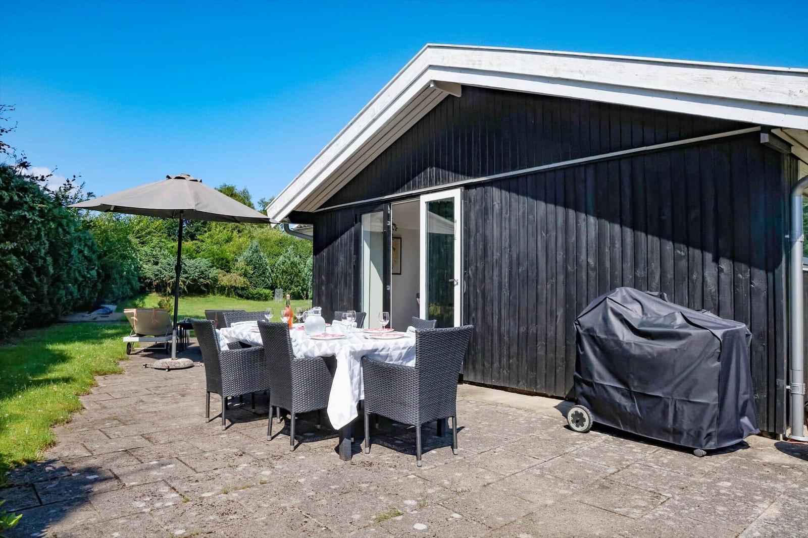 Außenbereich mit Tisch, Stühlen, Sonnenschirm und Grill. Hintergrund: grüner Garten.
