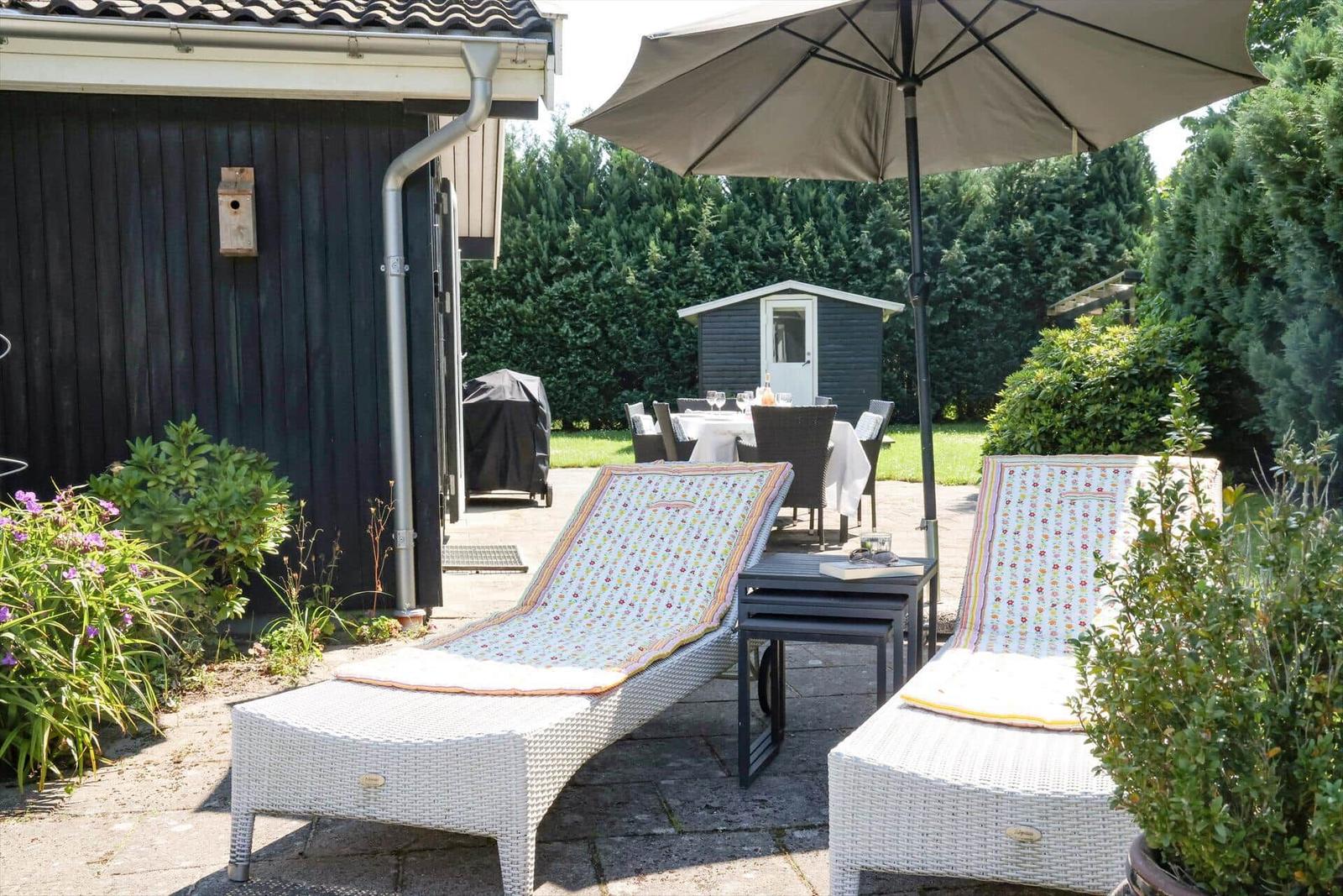 Terrasse mit Liegestühlen, Sonnenschirm und Tisch. Hintergrund: Grünfläche und Gartenhaus.